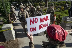 Protester holding No War on Iran sign during US Iran conflict demonstration as armed soldiers walk nearby