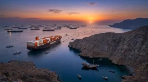 A high-angle drone landscape shot taken at dawn, capturing the rugged limestone cliffs of the Musandam peninsula, Oman, dropping into the deep blue sea at the entrance of the Strait of Hormuz. A large, orange Hapag-Lloyd container ship, with its hull featuring the distinctive orange 'H' logo on a white superstructure and stacked with orange and white containers, is positioned just at the entrance, appearing to hold position rather than actively transiting. Small naval patrol boats, with Omani markings and visible flags, create a tense, guarded line near the ship, suggesting a complex maritime traffic separation scheme and the potential mine threat. Further out, a massive cluster of hundreds of assorted cargo vessels, including numerous oil tankers and bulk carriers, are gathered on the other side of the strait, just within the Gulf, creating a vast, slightly chaotic-looking, clustered anchorage. The sunrise casts deep orange, purple, and blue hues across the sky and water, illuminating the scene and creating a sense of immense, stalled global trade on a grand natural stage, emphasizing the high-quality graphic request.