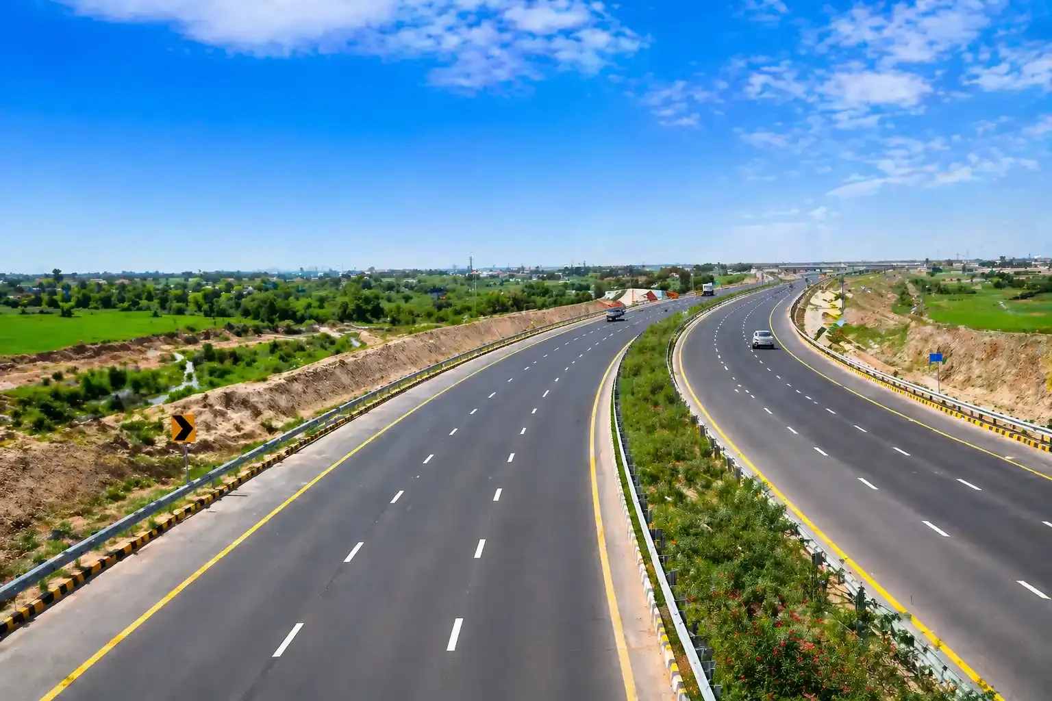 "Wide view of a modern six-lane motorway surrounded by green fields, with clear skies and well-maintained road infrastructure.