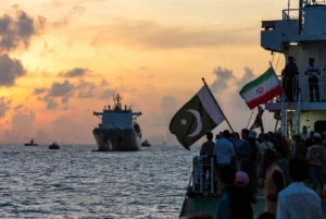 Pakistan seafarers standing on the deck of a seized vessel near Singapore waters at dramatic sunset with Pakistani and Iranian flags waving, representing safe return efforts by DPM Ishaq Dar