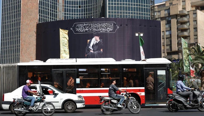 A busy street scene in Tehran, Iran, with a red and white city bus, motorbikes, and cars in the foreground, against a massive black religious billboard featuring Ayatollah Ali Khamenei and Arabic calligraphy.