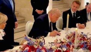 Donald Trump leaning forward to sign a document at a decorated formal dinner table, while French President Emmanuel Macron and Brigitte Macron look on.