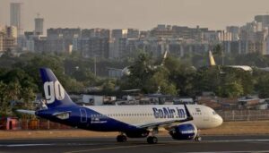 A blue and white GoAir passenger airplane taxiing on an airport runway with trees and a city skyline building background in India.
