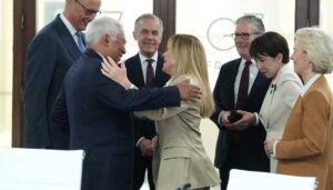 Italian Prime Minister Giorgia Meloni hugging António Costa at a G7 summit meeting, with UK Prime Minister Keir Starmer, Ursula von der Leyen, and other world leaders smiling in the background.