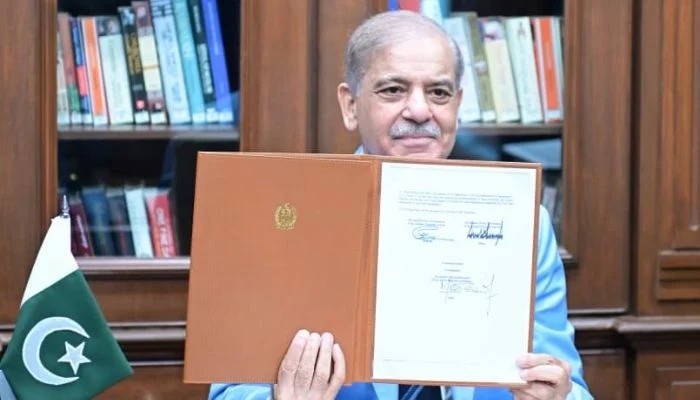 Prime Minister of Pakistan Shehbaz Sharif smiling and holding an official signed document folder next to the Pakistani flag.