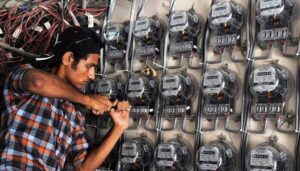 A young electrician or technician in a checkered shirt working on a wall covered with multiple digital and analog electricity meters, fixing wires with a tool