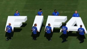 Workers in blue shirts carry giant white FIFA letters across a green football pitch during FIFA stake sale discussions