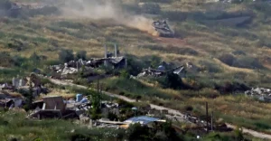 An armored main battle tank moves through a severely damaged, war-torn hillside village, illustrating the devastating impact of the ongoing US Iran conflict.