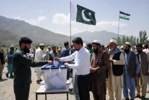 Voter casting ballot into box during AJK elections at outdoor polling station with Pakistani and AJK flags against mountain backdrop