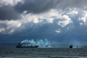 Cargo ships sailing under stormy skies in the Black Sea with smoke rising from one vessel after recent Black Sea attacks on civilian shipping