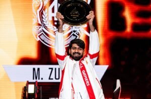 A smiling esports champion wearing a white and red All Gamers jacket, proudly lifting a gold championship plate trophy above his head on stage with a brightly lit orange background.