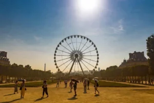 Tourists holding umbrellas for shade under intense sun near a large Ferris wheel in the Tuileries Garden during France hottest July heatwave in Paris