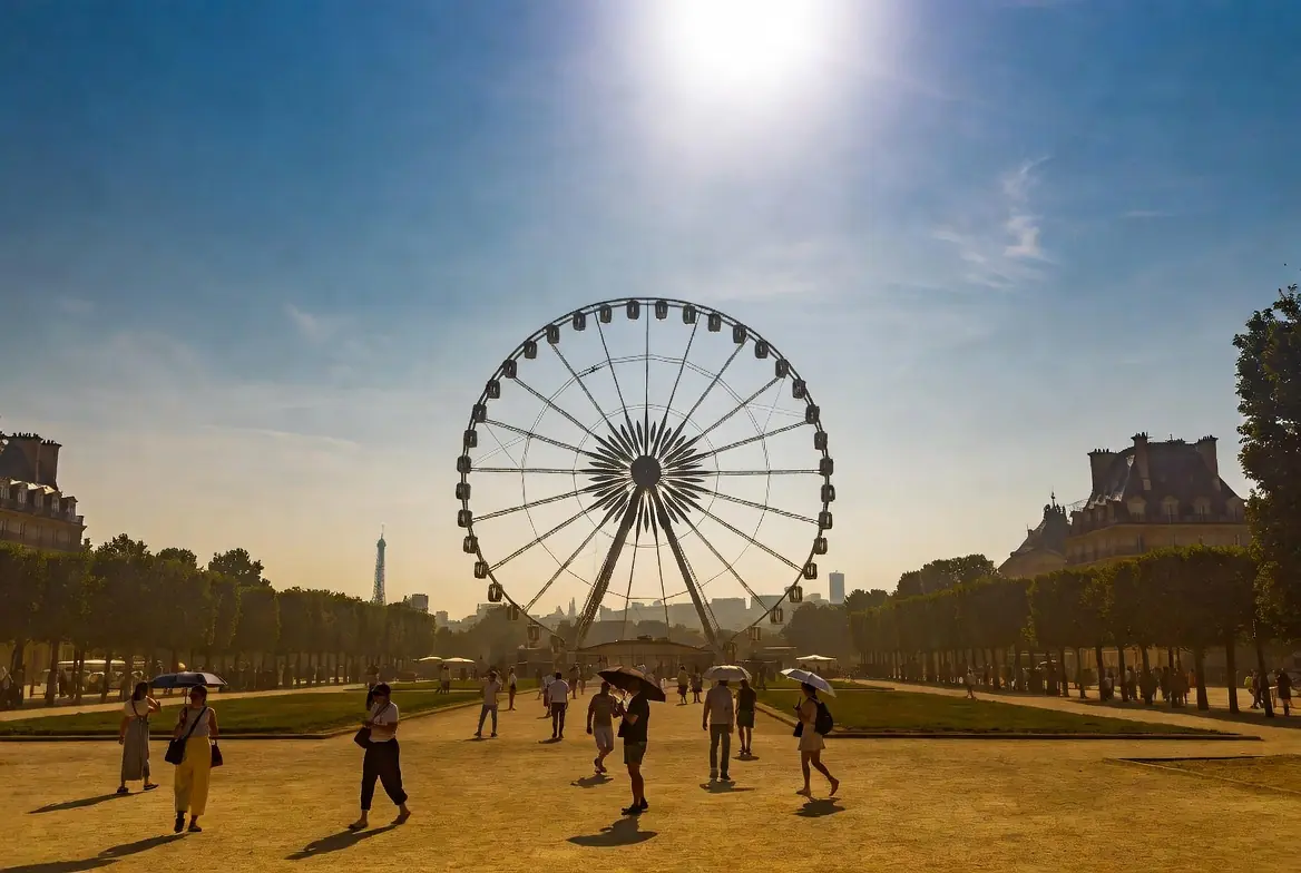 Tourists holding umbrellas for shade under intense sun near a large Ferris wheel in the Tuileries Garden during France hottest July heatwave in Paris
