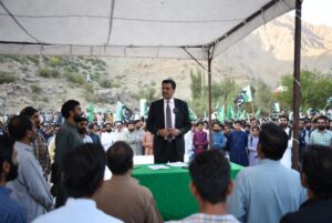 State Minister Bilal Azhar Kayani addressing supporters at outdoor corner meeting during PML-N AJK polls campaign in Muzaffarabad Division with green flags and mountains